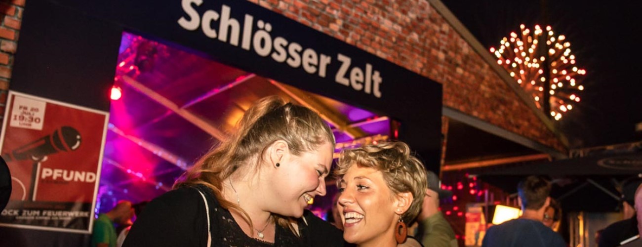 Zwei Frauen lachen und unterhalten sich vor einem beleuchteten Festzelt.