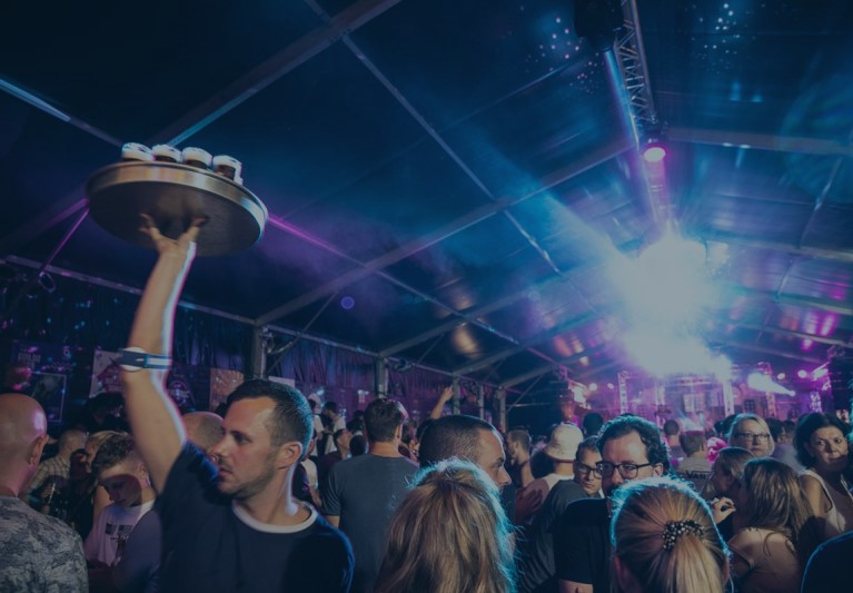 Eine Hand hält ein Tablett mit Gläsern hoch vor einer beleuchteten Bühne.